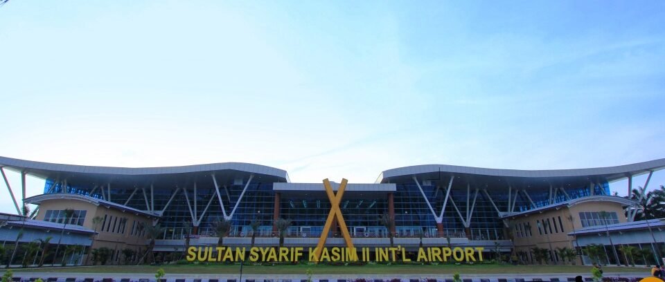 Sultan Syarif Kasim II International Airport terminal with a large yellow sign reading the airport name in front of glass façade.