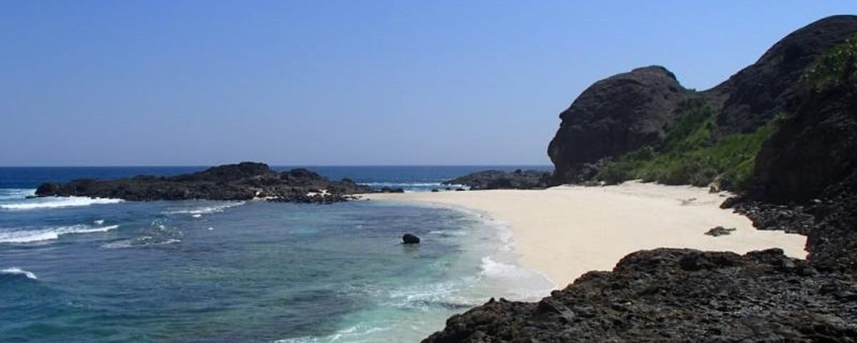 Sunny tropical beach with white sand, turquoise water, and dark rocky cliffs under a clear blue sky.