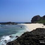 Sunny tropical beach with white sand, turquoise water, and dark rocky cliffs under a clear blue sky.