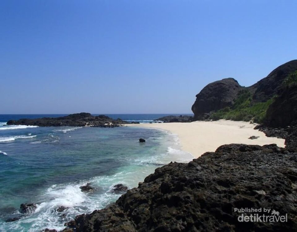 Sunny tropical beach with white sand, turquoise water, and dark rocky cliffs under a clear blue sky.