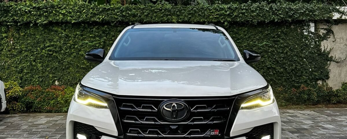 Front view of a white Toyota SUV parked on a brick driveway with green hedges and tropical plants in the background.