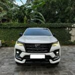 Front view of a white Toyota SUV parked on a brick driveway with green hedges and tropical plants in the background.