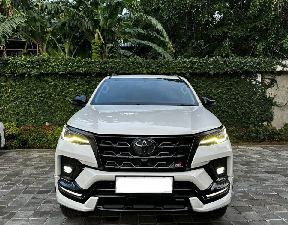 Front view of a white Toyota SUV parked on a brick driveway with green hedges and tropical plants in the background.