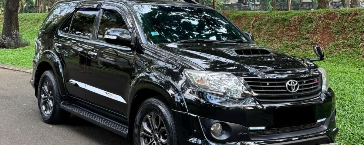 Black Toyota SUV parked on a tree-lined street with green grass nearby, reflecting surrounding foliage in its glossy surface.