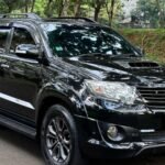 Black Toyota SUV parked on a tree-lined street with green grass nearby, reflecting surrounding foliage in its glossy surface.