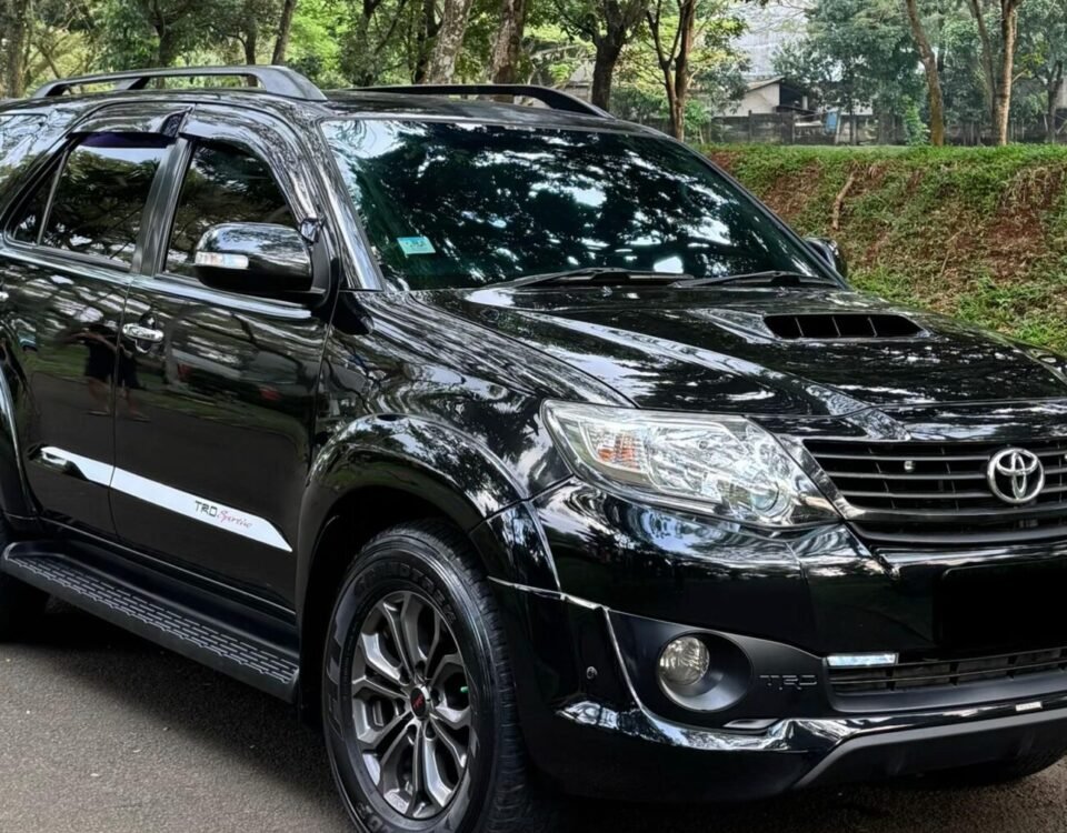 Black Toyota SUV parked on a tree-lined street with green grass nearby, reflecting surrounding foliage in its glossy surface.