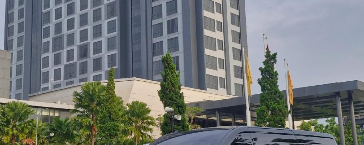 Exterior view of the Grand Mercure hotel with a tall blue-gray façade; a dark van is parked in the foreground and palm trees line the driveway.