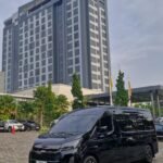 TikVideo.App_7584053555082923285_1 - Dyantransport Exterior view of the Grand Mercure hotel with a tall blue-gray façade; a dark van is parked in the foreground and palm trees line the driveway.