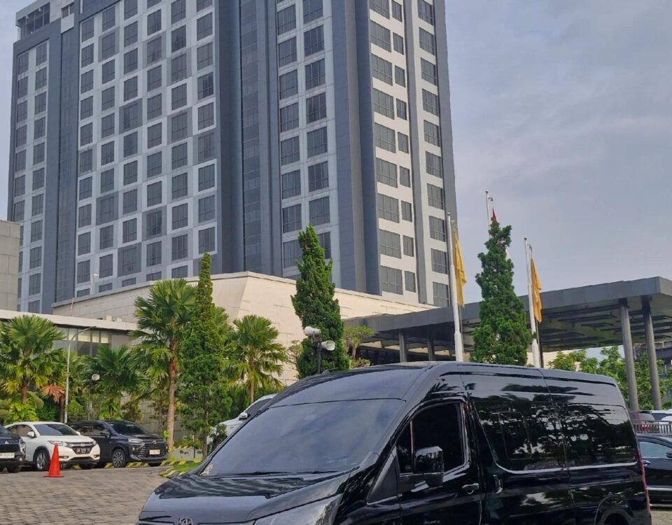 Exterior view of the Grand Mercure hotel with a tall blue-gray façade; a dark van is parked in the foreground and palm trees line the driveway.