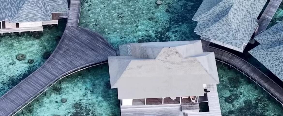 Aerial view of overwater bungalows connected by wooden walkways over clear turquoise water with a coral reef below.