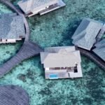 Aerial view of overwater bungalows connected by wooden walkways over clear turquoise water with a coral reef below.