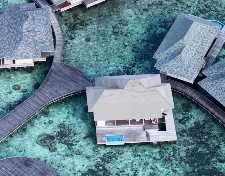 Aerial view of overwater bungalows connected by wooden walkways over clear turquoise water with a coral reef below.