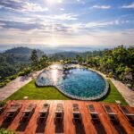 Sunlit hilltop resort with a kidney-shaped pool, wooden deck, and row of lounge chairs overlooking distant mountains and sea.