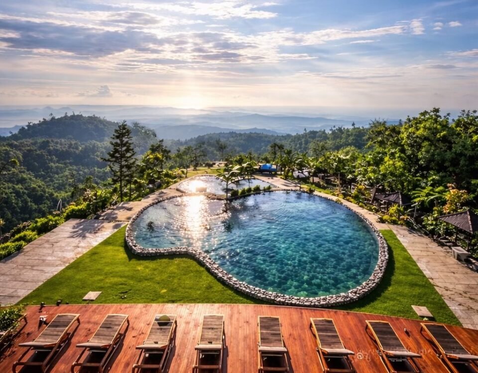 Sunlit hilltop resort with a kidney-shaped pool, wooden deck, and row of lounge chairs overlooking distant mountains and sea.