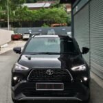 Front view of a black Toyota SUV parked in a narrow alley, raindrops on the hood, with gray metal storefront shutters on the right and green trees in the background.
