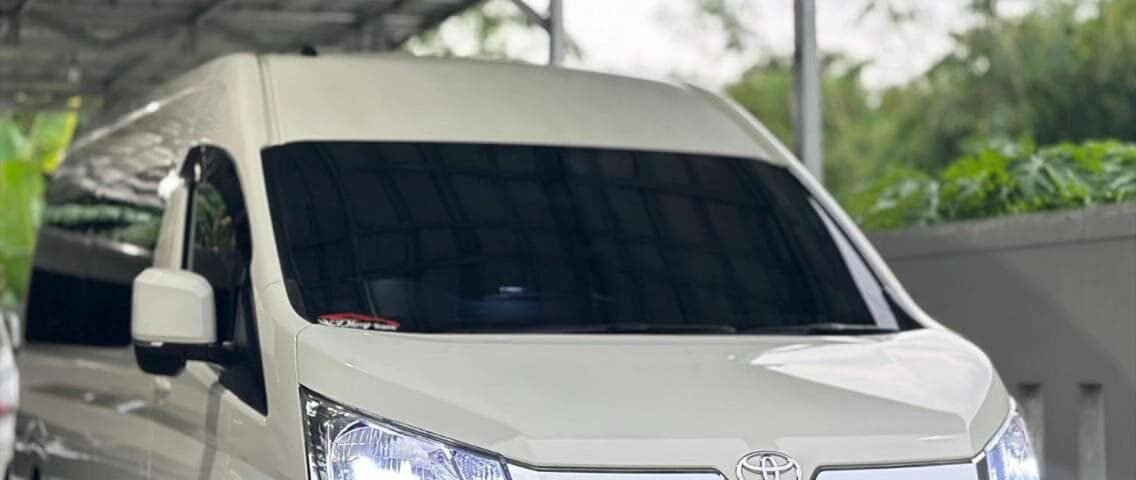 White Toyota van parked under a metal carport, facing slightly to the right, headlights on.