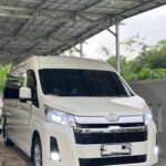 White Toyota van parked under a metal carport, facing slightly to the right, headlights on.