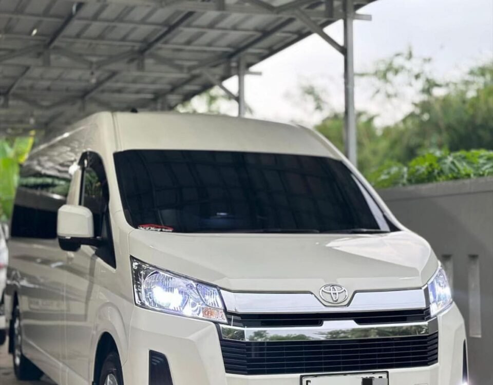 White Toyota van parked under a metal carport, facing slightly to the right, headlights on.