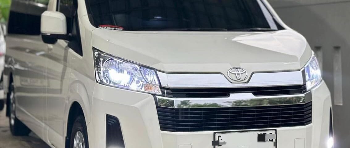 White Toyota van parked under a metal carport, facing slightly to the right, headlights on.