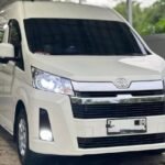 White Toyota van parked under a metal carport, facing slightly to the right, headlights on.