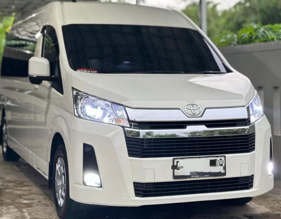White Toyota van parked under a metal carport, facing slightly to the right, headlights on.