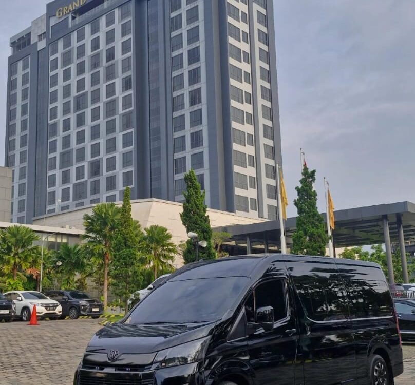 Black Toyota high-roof passenger van parked on a brick-paved lot with palm trees in the background. The windows are tinted and the vehicle has a glossy finish.
