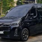Black Toyota high-roof passenger van parked on a brick-paved lot with palm trees in the background. The windows are tinted and the vehicle has a glossy finish.