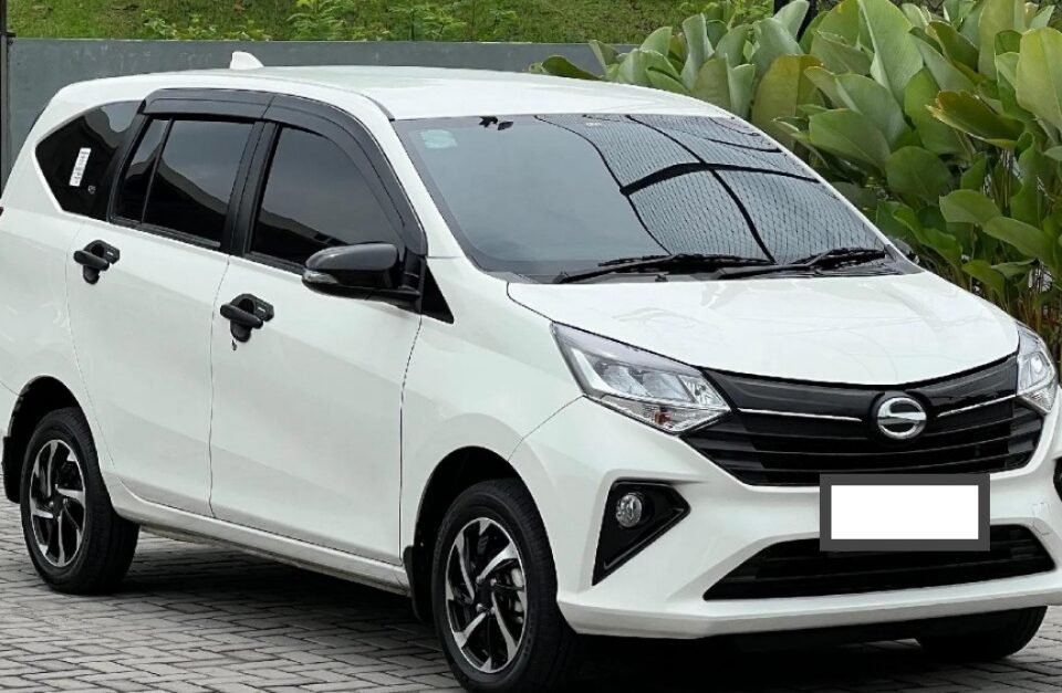 White compact MPV parked on a brick-paved parking lot beside green shrubs and a chain-link fence, shown in a front-three-quarter view.