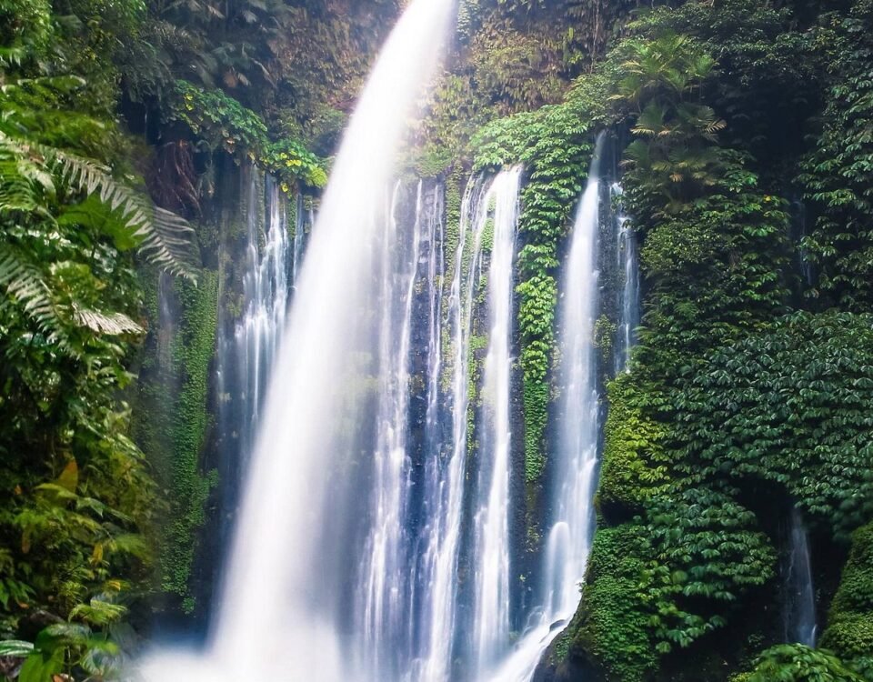 Waterfall pouring over a lush, mossy cliff into a rocky pool, surrounded by dense tropical jungle greenery