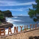 Coastal overlook with a rocky shore, blue water, and orange letters spelling 'BANYU TIBO' along a stone path.