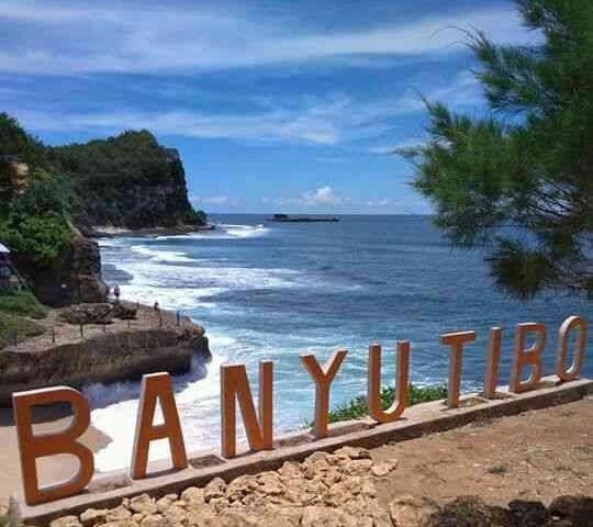 Coastal overlook with a rocky shore, blue water, and orange letters spelling 'BANYU TIBO' along a stone path.