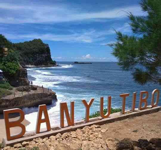Coastal overlook with a rocky shore, blue water, and orange letters spelling 'BANYU TIBO' along a stone path.