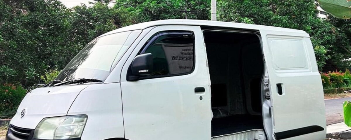 White cargo/van with its side door open, parked on a street with green plants and trees in the background.
