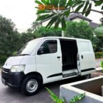White cargo/van with its side door open, parked on a street with green plants and trees in the background.