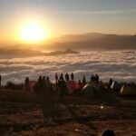 Sunrise over a cloud-filled valley seen from a hilltop campsite; colorful tents and silhouetted campers gather at the ridge.