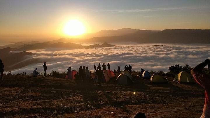 Sunrise over a cloud-filled valley seen from a hilltop campsite; colorful tents and silhouetted campers gather at the ridge.