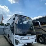bus - Dyantransport Front three-quarter view of a white tour bus with a large windshield, parked on a gravel lot beside another bus under a cloudy blue sky.