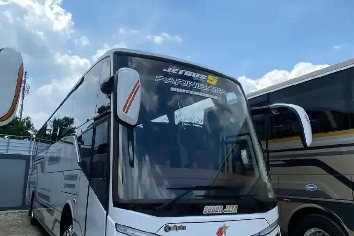 Front three-quarter view of a white tour bus with a large windshield, parked on a gravel lot beside another bus under a cloudy blue sky.