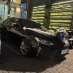 Black wedding car with white ribbons and a large floral bouquet on the hood, parked outside a lit venue with a green wall of plants behind it.