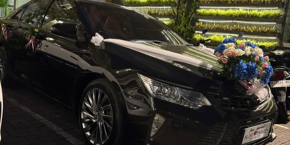 Black wedding car with white ribbons and a large floral bouquet on the hood, parked outside a lit venue with a green wall of plants behind it.