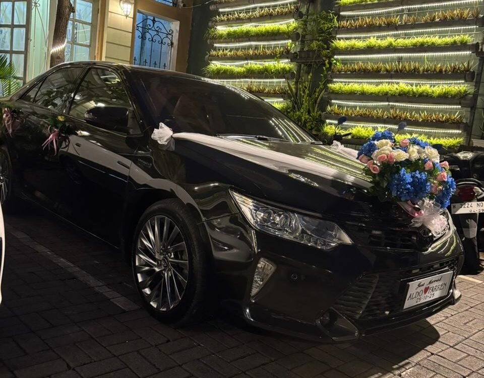 Black wedding car with white ribbons and a large floral bouquet on the hood, parked outside a lit venue with a green wall of plants behind it.