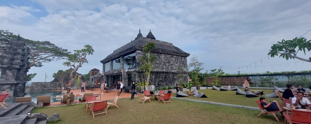 Stone pavilion and historic stone building on a grassy terrace with outdoor seating and string lights, visitors walking around.