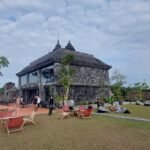 Stone pavilion and historic stone building on a grassy terrace with outdoor seating and string lights, visitors walking around.