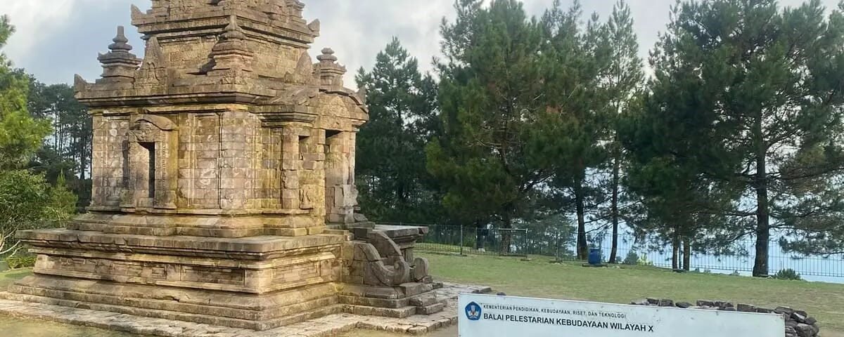 Stone ancient temple ruin (Candi Gedong II) with a multi-tiered stone structure set on a grassy area, surrounded by trees and a low fence nearby.
