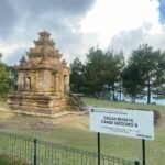 Stone ancient temple ruin (Candi Gedong II) with a multi-tiered stone structure set on a grassy area, surrounded by trees and a low fence nearby.