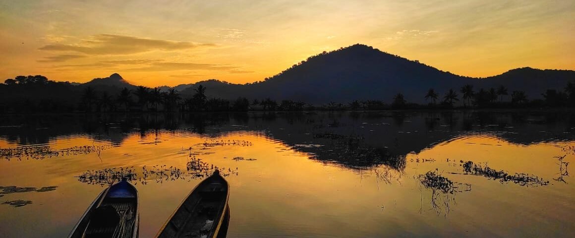 Two small wooden boats float on a calm lake at sunset, with palm silhouettes and a hill in the distance, water reflecting orange and gold hues.