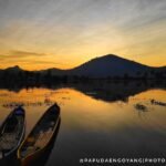 Two small wooden boats float on a calm lake at sunset, with palm silhouettes and a hill in the distance, water reflecting orange and gold hues.