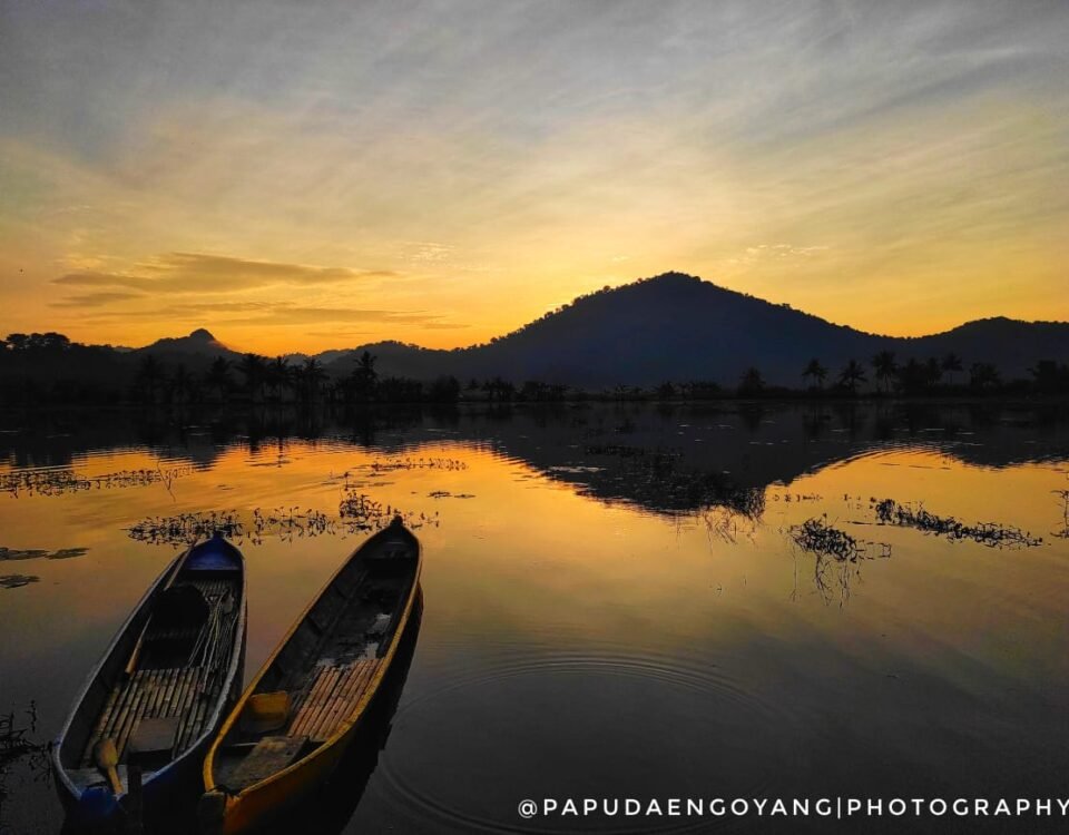 Two small wooden boats float on a calm lake at sunset, with palm silhouettes and a hill in the distance, water reflecting orange and gold hues.