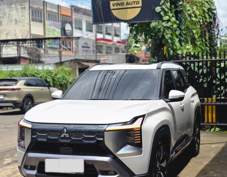 White Mitsubishi SUV parked on a street with a dark blue VINS AUTO sign overhead, vines climbing around the sign and utility lines above.