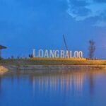 Lakeside park at dusk with a large sign reading 'LOANG BALOO', illuminated by street lamps and flanked by two gazebos overlooking calm water.